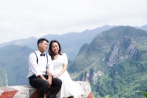A couple is seated together on a stone platform with a mountainous landscape in the background. The man is wearing a dress shirt with a bow tie and suspenders, while the woman is in a white dress. The scene conveys a sense of serenity and romance amidst the lush greenery of the mountains.
