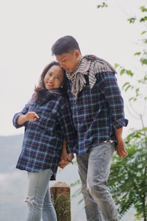 A couple stands hand in hand wearing matching plaid shirts and distressed jeans. Both are smiling warmly at each other, with a serene outdoor background featuring greenery. The man has a scarf draped around his neck and leans slightly towards the woman, creating a warm, affectionate pose.