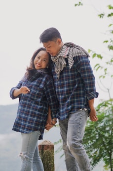 A couple stands hand in hand wearing matching plaid shirts and distressed jeans. Both are smiling warmly at each other, with a serene outdoor background featuring greenery. The man has a scarf draped around his neck and leans slightly towards the woman, creating a warm, affectionate pose.
