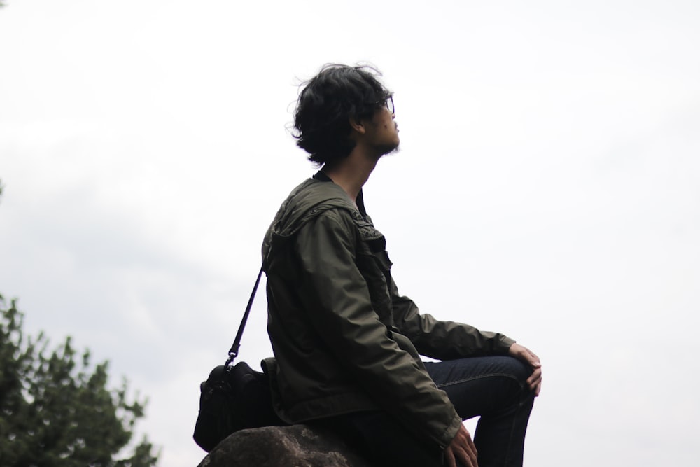 man in black jacket and blue denim jeans sitting on rock