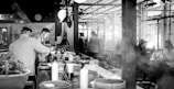 In a bustling kitchen setting, two chefs are wearing uniforms and hats, diligently preparing food amidst various kitchen tools and ingredients. The atmosphere appears busy and professional, with plates and bottles arranged on the counter. Diners are visible in the background enjoying their meals at tables in a restaurant, adding to the lively ambiance.