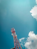 A red and white telecommunications tower extends upwards against a backdrop of a clear blue sky. Fluffy white clouds drift nearby, adding contrast to the expansive sky. The structure of the tower is detailed with metal beams and several antennae.
