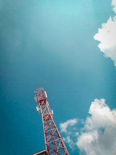 A red and white telecommunications tower extends upwards against a backdrop of a clear blue sky. Fluffy white clouds drift nearby, adding contrast to the expansive sky. The structure of the tower is detailed with metal beams and several antennae.