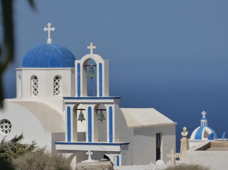 A picturesque scene featuring a traditional Greek church with a blue dome and white walls. The bell tower is prominent with multiple bells and crosses. In the background, the deep blue color of the sea contrasts with the clear sky.