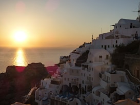 The image captures a stunning sunset over a coastal village with white-washed buildings. The sun is low on the horizon, casting a golden glow across the sea. The buildings are stacked on a hillside, demonstrating classic Mediterranean architecture.
