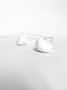 A pair of white wired earphones seen in close-up on a smooth, pale surface. The earphones are symmetrically aligned, with one slightly in front of the other, both showcasing their design and detailing.