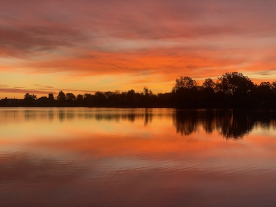 A tranquil lake reflects the vibrant colors of a sunset with an orange and pink sky above. The silhouette of trees lines the horizon, creating a peaceful and serene landscape.