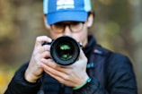 A person wearing a blue cap and glasses is holding a camera with a large lens, focusing intently on capturing a photograph. The background is softly blurred, drawing attention to the camera and the person.