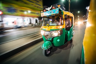 A green and yellow auto rickshaw with passengers is moving swiftly down a street at night, with blurred shops and streetlights in the background indicating the vehicle's speed.
