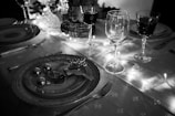 A beautifully set dining table features festive decorations, including a plate adorned with ornate table settings like a glittering reindeer and small baubles. Several wine glasses are elegantly arranged, while warm string lights illuminate the scene, casting a soft glow.