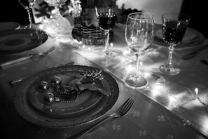A beautifully set dining table features festive decorations, including a plate adorned with ornate table settings like a glittering reindeer and small baubles. Several wine glasses are elegantly arranged, while warm string lights illuminate the scene, casting a soft glow.