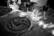 A beautifully set dining table features festive decorations, including a plate adorned with ornate table settings like a glittering reindeer and small baubles. Several wine glasses are elegantly arranged, while warm string lights illuminate the scene, casting a soft glow.