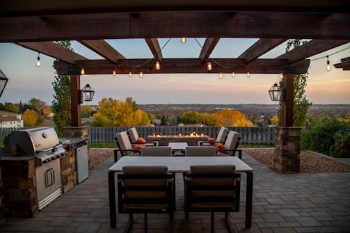 A patio with a pergola, featuring string lights and a cozy outdoor dining area with cushioned chairs. The patio includes a built-in grill on the left and a fire pit in the center. The setting overlooks a serene landscape with autumn foliage in the background under a soft, evening sky.