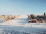 A snowy landscape with a ski slope and surrounding frost-covered trees. A building is positioned at the base of the slope, and ski lifts are visible climbing up the hill. The sky is clear and blue, adding contrast to the white scenery.