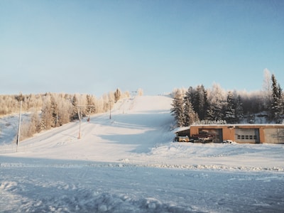 A snowy landscape with a ski slope and surrounding frost-covered trees. A building is positioned at the base of the slope, and ski lifts are visible climbing up the hill. The sky is clear and blue, adding contrast to the white scenery.