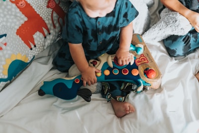 A toddler sits on a bed playing with a colorful guitar-shaped toy. The toy features bright buttons and interactive elements. The child is wearing a dark teal outfit and is surrounded by playful, animal-themed bedding with images of llamas and monkeys.