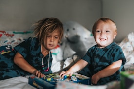 Two children are sitting together on a bed, engaged with a colorful book or toy. One child has longish hair and wears a beaded necklace, while the other is smiling brightly. In the background, there are plush toys and patterned bed sheets.