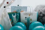 A festive arrangement featuring gift bags surrounded by blue balloons. One bag prominently displays a large number '1', embellished with playful animal illustrations, suggesting a first birthday celebration. The setup includes a cozy, furry blanket and decorative lights, adding a warm, inviting touch.