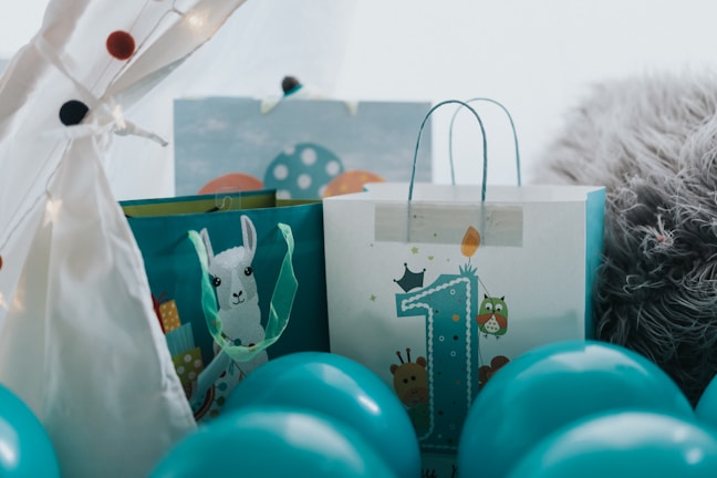 A festive arrangement featuring gift bags surrounded by blue balloons. One bag prominently displays a large number '1', embellished with playful animal illustrations, suggesting a first birthday celebration. The setup includes a cozy, furry blanket and decorative lights, adding a warm, inviting touch.