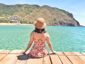 woman in red and white floral spaghetti strap dress sitting on brown wooden dock during daytime