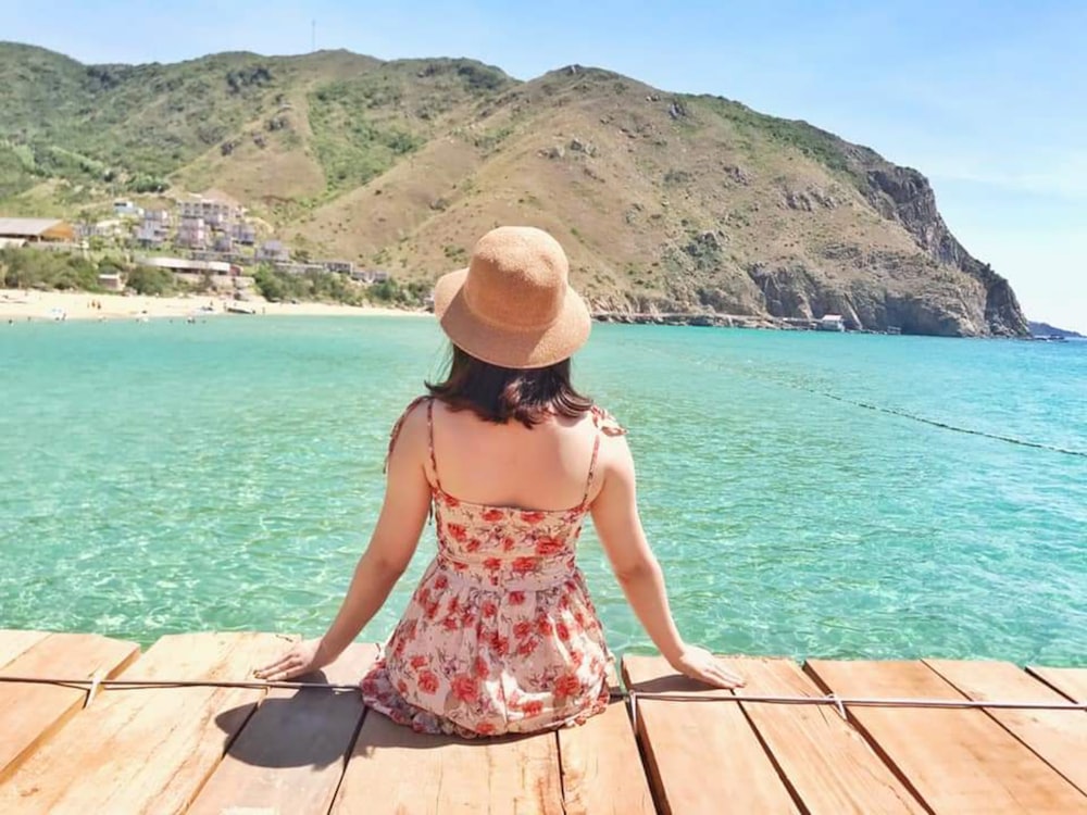 woman in red and white floral spaghetti strap dress sitting on brown wooden dock during daytime