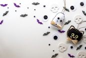 A playful arrangement of Halloween-themed decorations including felt crafts of a ghost and a black cat, surrounded by various sizes of googly eyes, small bats with cartoonish features, sparkly purple bat cutouts, and flat skull shapes on a light background.