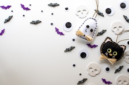 A playful arrangement of Halloween-themed decorations including felt crafts of a ghost and a black cat, surrounded by various sizes of googly eyes, small bats with cartoonish features, sparkly purple bat cutouts, and flat skull shapes on a light background.