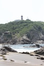 A picturesque scene featuring a sandy beach with large rocks in the foreground and waves gently crashing onto the shore. Behind the beach, a steep, green hillside rises, topped with a red and white striped lighthouse and a small building. The sky is clear and bright.