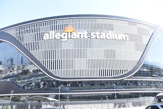 A large, modern stadium with reflective glass surfaces and sleek architectural lines. The name 'Allegiant Stadium' is prominently displayed in white letters, accompanied by a sun logo. Surrounding the stadium are various parked vehicles, visible through the reflection on the glass.