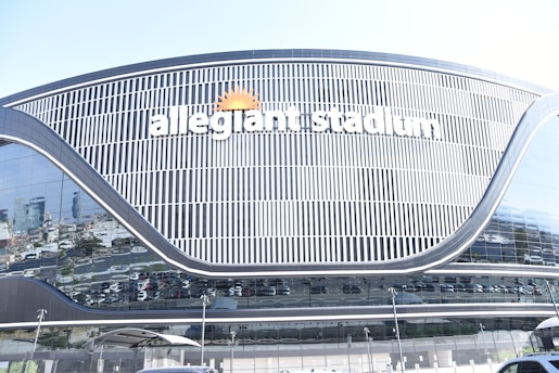 A large, modern stadium with reflective glass surfaces and sleek architectural lines. The name 'Allegiant Stadium' is prominently displayed in white letters, accompanied by a sun logo. Surrounding the stadium are various parked vehicles, visible through the reflection on the glass.