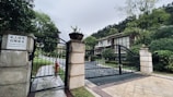 A gated entrance to a private property, featuring ornate black metal gates with stone pillars. A sign displays text in both Chinese and English, indicating restricted access for villa guests only. Lush greenery and trees surround the area. The driveway leads up to a modern villa with large windows nestled among the trees.