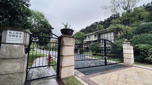 A gated entrance to a private property, featuring ornate black metal gates with stone pillars. A sign displays text in both Chinese and English, indicating restricted access for villa guests only. Lush greenery and trees surround the area. The driveway leads up to a modern villa with large windows nestled among the trees.