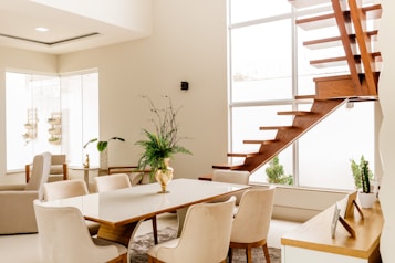 A modern, minimalist interior space featuring a wooden staircase leading to an upper level. Light beige walls and large windows allow natural light to fill the room. The center of the area has a white dining table with light beige upholstered chairs around it. Several potted plants, including a prominent fern in a gold vase on the table, add a touch of greenery. A soft, plush rug is under the dining table, and a sleek sideboard with decorative items is positioned nearby. The clean lines and neutral tones create an open and airy ambiance.