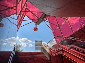 An urban stairway and escalator leads up to an exit that opens to a bright blue sky adorned with scattered clouds. The structure is framed by vibrant red and pink glass panels, creating a modern, geometric aesthetic. A round sign with an 'M' hangs prominently near the top. Reflections in the glass enhance the colorful atmosphere.