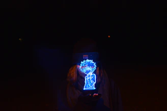 A glowing blue holographic figure is held by a person wearing a beanie and scarf, set against a dark background. The figure appears animated and is the central focus, emitting a bright light that contrasts with the surrounding darkness.