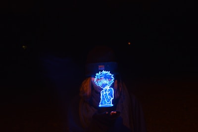 A glowing blue holographic figure is held by a person wearing a beanie and scarf, set against a dark background. The figure appears animated and is the central focus, emitting a bright light that contrasts with the surrounding darkness.