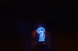 A glowing blue holographic figure is held by a person wearing a beanie and scarf, set against a dark background. The figure appears animated and is the central focus, emitting a bright light that contrasts with the surrounding darkness.
