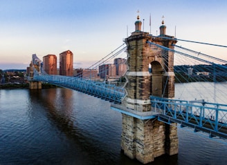 A suspension bridge crosses over a river, connecting urban landscapes on both sides. The bridge features prominent stone towers and sky-blue cables. In the background, modern high-rise buildings with reflective glass catch the light of the setting sun, which gives the scene a warm glow. The river beneath reflects the colors of the sky and surrounding structures.