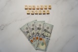 A marble surface displays five 100-dollar bills arranged in a fan shape. Above the bills, the words PASSIVE INCOME are spelled using wooden Scrabble tiles.