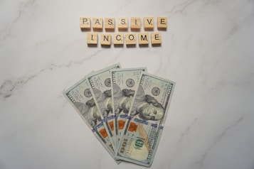 A marble surface displays five 100-dollar bills arranged in a fan shape. Above the bills, the words PASSIVE INCOME are spelled using wooden Scrabble tiles.