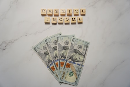 A marble surface displays five 100-dollar bills arranged in a fan shape. Above the bills, the words PASSIVE INCOME are spelled using wooden Scrabble tiles.