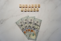 A marble surface displays five 100-dollar bills arranged in a fan shape. Above the bills, the words PASSIVE INCOME are spelled using wooden Scrabble tiles.