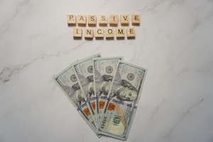 A marble surface displays five 100-dollar bills arranged in a fan shape. Above the bills, the words PASSIVE INCOME are spelled using wooden Scrabble tiles.