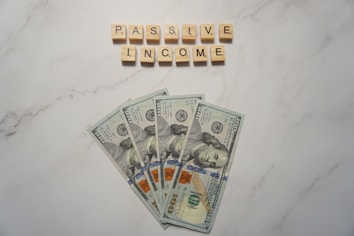 A marble surface displays five 100-dollar bills arranged in a fan shape. Above the bills, the words PASSIVE INCOME are spelled using wooden Scrabble tiles.