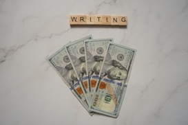 Five one-hundred dollar bills are fanned out on a marble-like surface, with wooden letter tiles spelling 'WRITING' arranged above them.