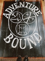 A wooden floor features a bold white design with the words 'Adventure Bound' around three circular illustrations. The illustrations depict a boot, a crossed paddle design, and a backpack. The floorboards are dark with a reddish-brown border, and a pair of shoes is visible at the bottom.