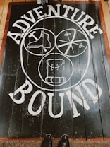 A wooden floor features a bold white design with the words 'Adventure Bound' around three circular illustrations. The illustrations depict a boot, a crossed paddle design, and a backpack. The floorboards are dark with a reddish-brown border, and a pair of shoes is visible at the bottom.