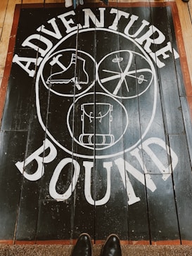 A wooden floor features a bold white design with the words 'Adventure Bound' around three circular illustrations. The illustrations depict a boot, a crossed paddle design, and a backpack. The floorboards are dark with a reddish-brown border, and a pair of shoes is visible at the bottom.