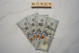 Five one-hundred dollar bills are fanned out on a marble surface. Above them, Scrabble tiles spell out the word 'MONEY'.