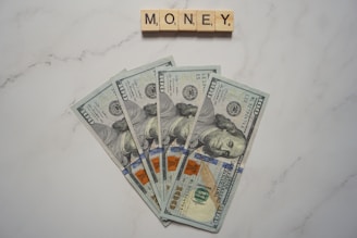 Five one-hundred dollar bills are fanned out on a marble surface. Above them, Scrabble tiles spell out the word 'MONEY'.